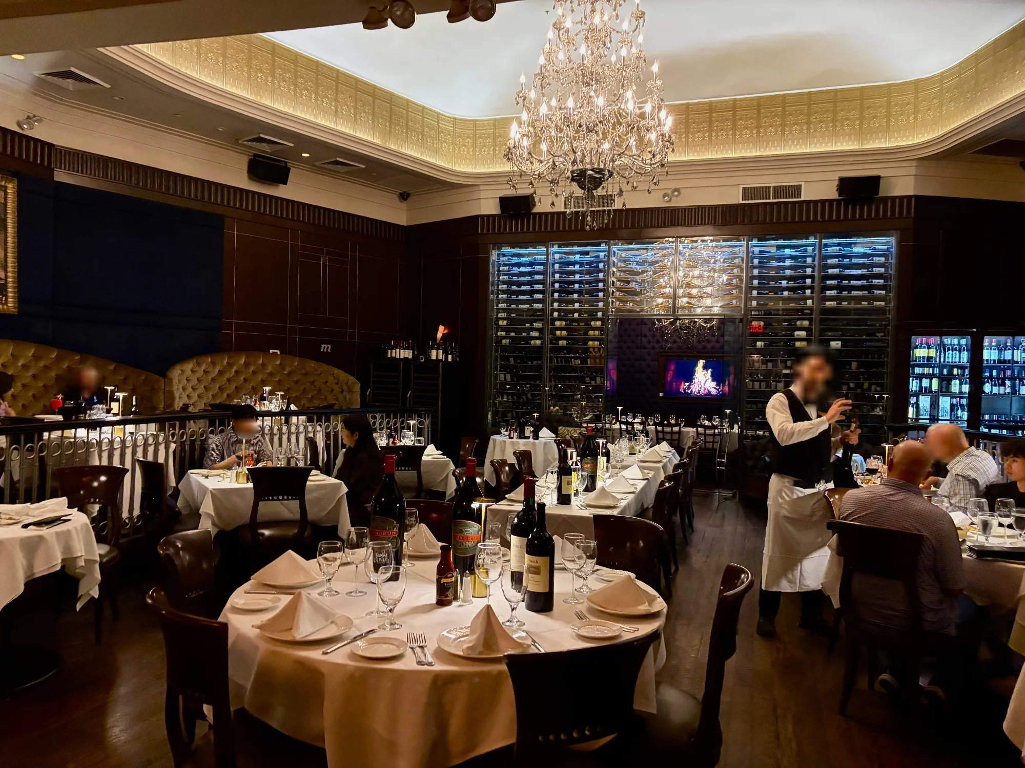 A dining room with white-tablecloth tables, high-backed booths, dark wood walls, a chandelier, wine racks, and diners.