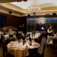 A dining room with white-tablecloth tables, high-backed booths, dark wood walls, a chandelier, wine racks, and diners.