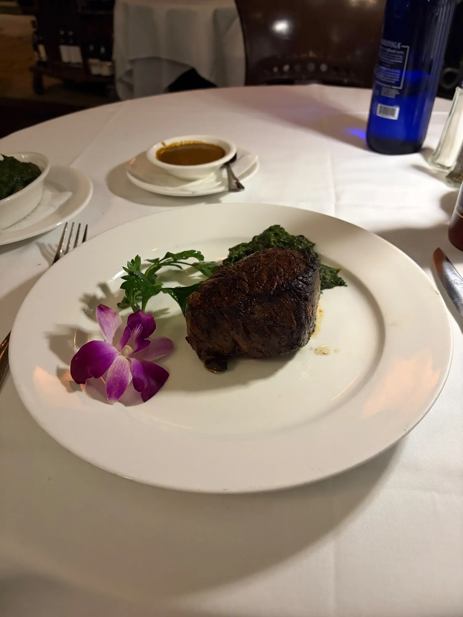A filet mignon on a white plate with greens, parsley, and a purple flower, with a sauce boat behind it.