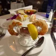 Four shrimp arranged around a metal cocktail bowl with sauce, lemon wedges, and a purple flower.