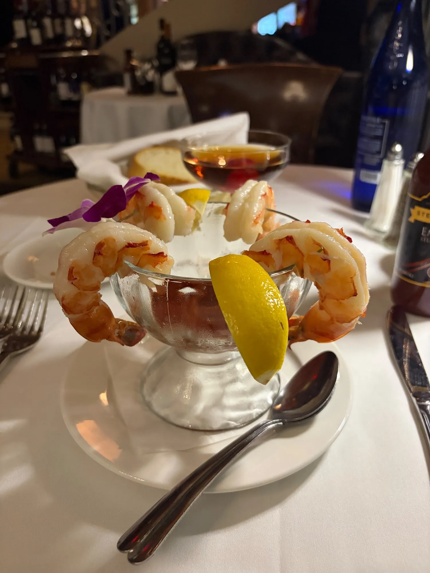 Four shrimp arranged around a metal cocktail bowl with sauce, lemon wedges, and a purple flower.