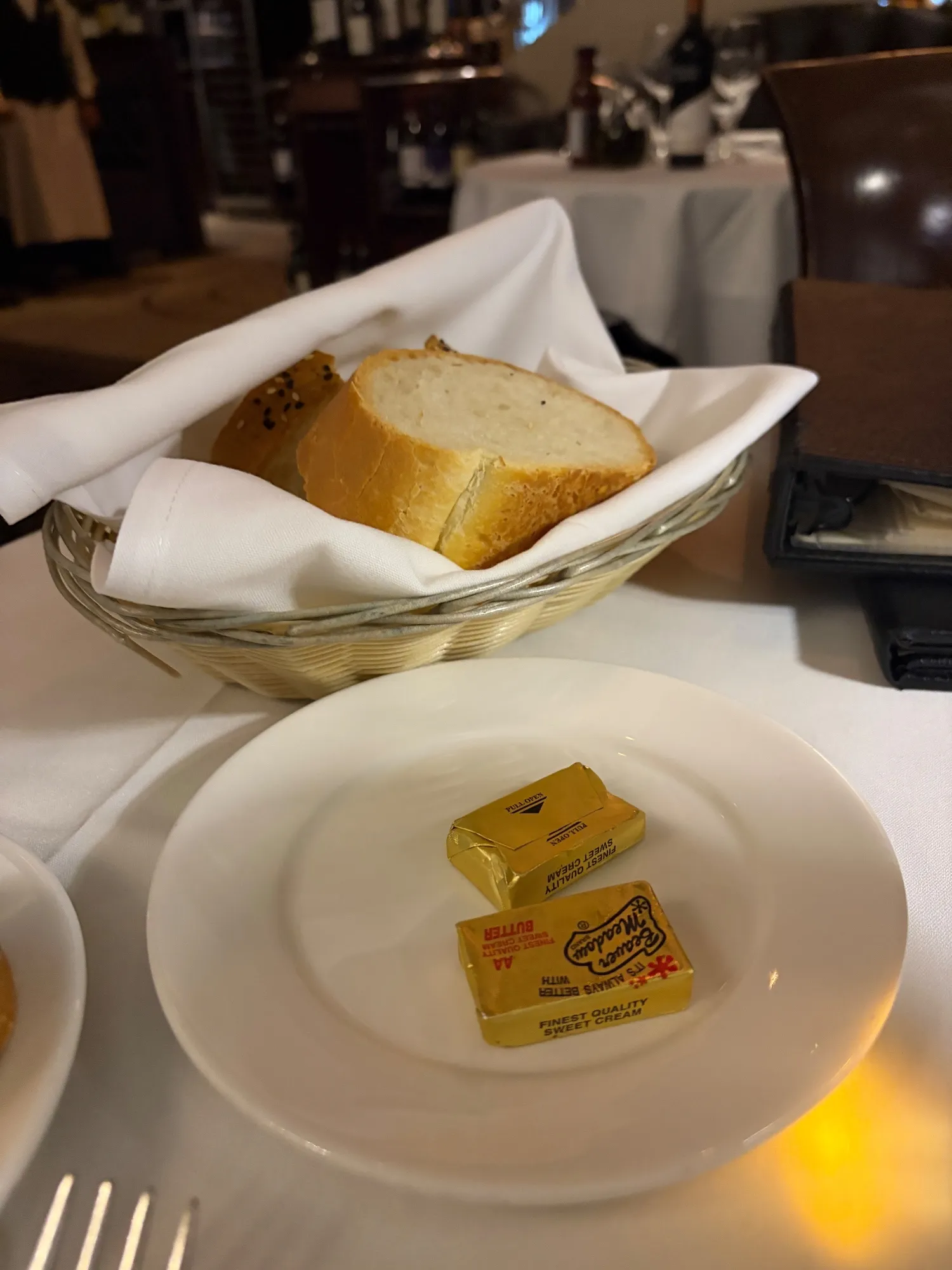 A cloth-lined basket of sliced bread beside two wrapped butter packets on a white plate.