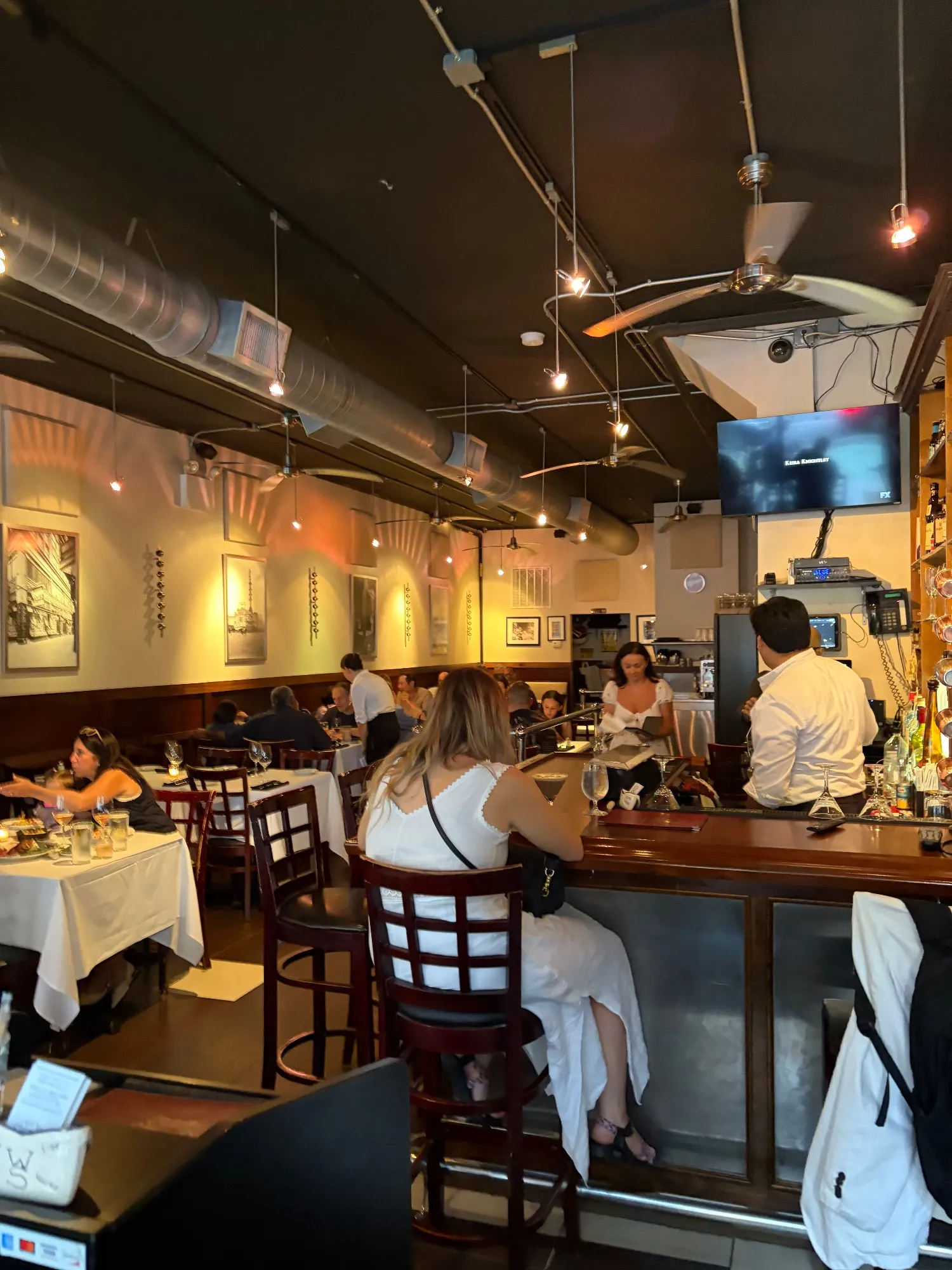 Interior dining room with white-clothed tables, a bar to the right, exposed ductwork and a wall-mounted TV; patrons seated throughout.
