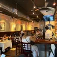 Interior dining room with white-clothed tables, a bar to the right, exposed ductwork and a wall-mounted TV; patrons seated throughout.