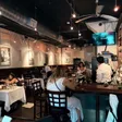 Interior dining room with white-clothed tables, a bar to the right, exposed ductwork and a wall-mounted TV; patrons seated throughout.