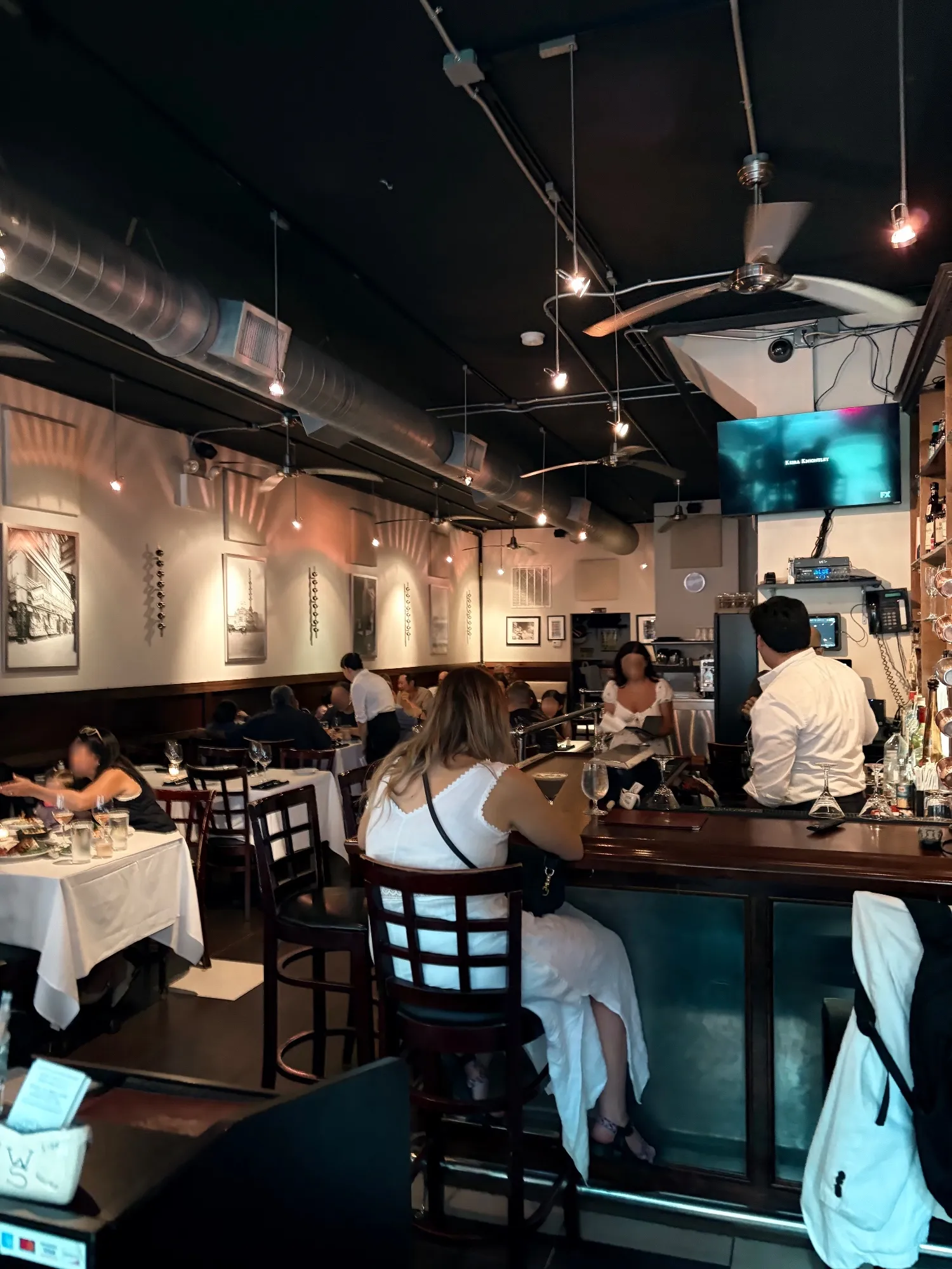 Interior dining room with white-clothed tables, a bar to the right, exposed ductwork and a wall-mounted TV; patrons seated throughout.